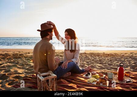 Frau, die mit Hut spielt, während sie beim Mann am Strand sitzt Stockfoto