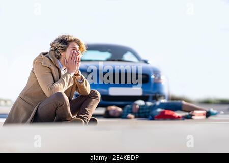 Verzweifelter junger Mann auf dem Boden mit Jungen im Hintergrund sitzen Mit dem Auto nach einem Unfall liegen Stockfoto