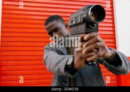 Junger Mann hält 1980s Filmkamera, während er gegen Rot steht Wand Stockfoto
