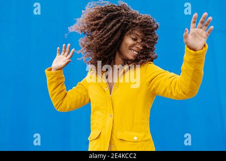 Lächelnde junge Frau in gelber Jacke wirft Haare im Stehen Gegen blaue Wand Stockfoto
