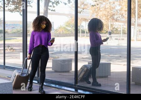 Junge Frau in schützende Gesichtsmaske mit Smartphone, während Spaziergang mit Gepäck am Glas Stockfoto