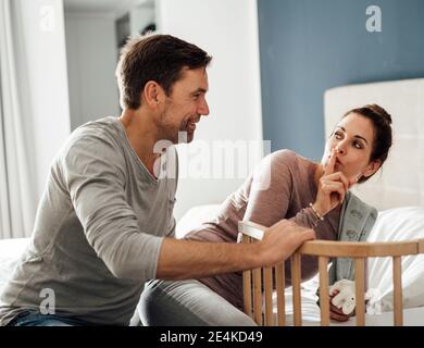 Reife Frau mit Finger auf den Lippen Blick auf Mann durch Kinderbett im Schlafzimmer Stockfoto