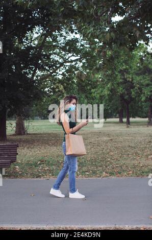 Junge Frau mit Papiertüte mit Handy beim Gehen Im öffentlichen Park während der COVID-19 Stockfoto