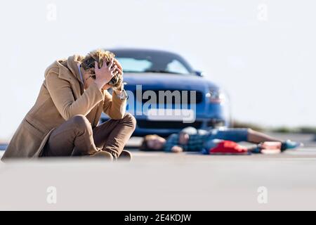 Verzweifelter junger Mann auf dem Boden mit Jungen im Hintergrund sitzen Mit dem Auto nach einem Unfall liegen Stockfoto