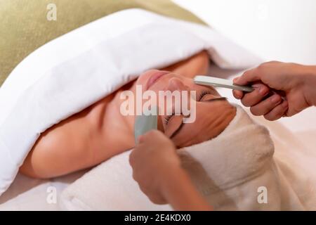 Kosmetikerin hält Gua Sha auf dem Gesicht der weiblichen Kundin bei der Gesundheit Spa Stockfoto