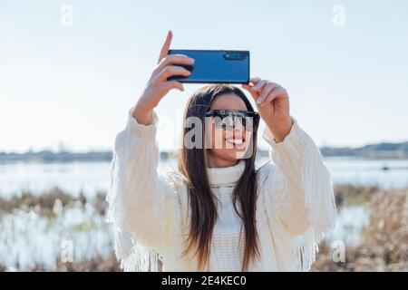 Schöne Frau trägt eine Sonnenbrille und nimmt Selfie mit einem Smartphone entgegen Der Himmel ist klar Stockfoto