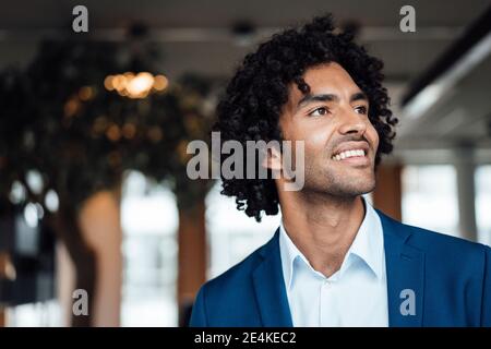 Lächelnder, hübscher junger Geschäftsmann mit lockigen schwarzen Haaren, die wegschauen Im Büro Stockfoto