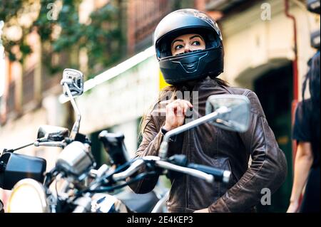 Weibliche Biker befestigen Sturzhelm, während sie auf dem Motorrad auf dem Bürgersteig sitzen Stockfoto