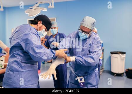 Assistentin hilft dem Arzt beim Tragen von Schutzhandschuhen im Stehen Operationsraum Stockfoto