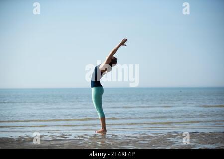 Reife Frau mit erhobenen Armen Training am Strand gegen klar Himmel Stockfoto