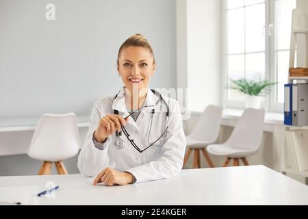 Glücklicher junger Arzt, der am Tisch in ihrem Büro sitzt, lächelt und auf die Kamera schaut Stockfoto
