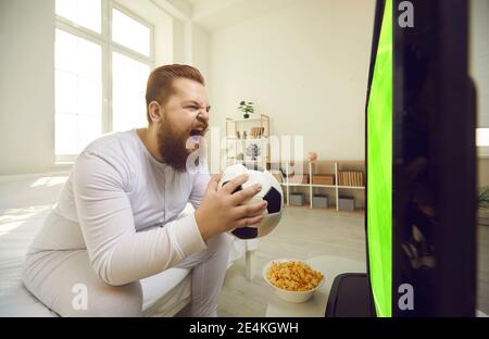 Lustige mollige Fan sitzt auf dem Bett in Pyjamas und beobachten Aufregendes Fußballspiel im Fernsehen Stockfoto