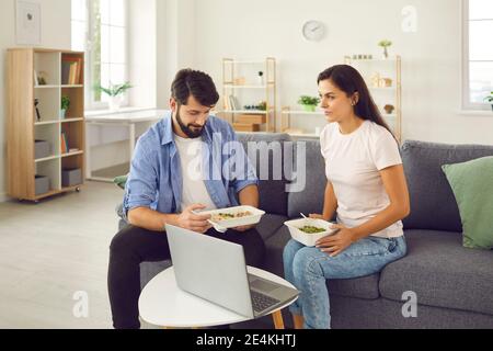 Mann und Frau sitzen zu Hause auf der Couch vor einem Laptop und essen Essen aus einer Lieferung. Stockfoto