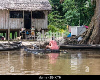 Amazonas-Fluss, Peru - 13. Mai 2016: Holzhaus auf Stelzen und eine Frau, die Wäsche auf einer hölzernen Plattform am Flussufer macht. Kleines Dorf am Amazonas Stockfoto