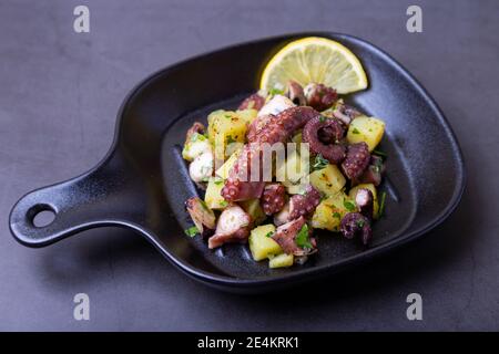 Warmer Salat mit Oktopus, Kartoffeln und Zitrone in einer schwarzen kleinen Pfanne. Nahaufnahme, schwarzer Hintergrund. Stockfoto