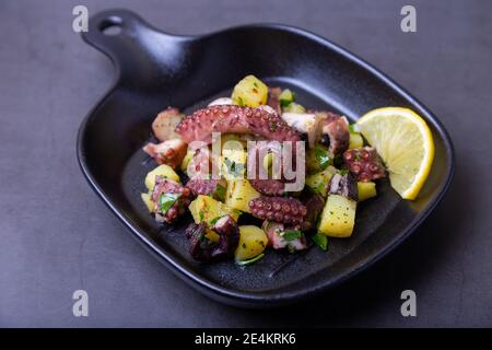 Warmer Salat mit Oktopus, Kartoffeln und Zitrone in einer schwarzen kleinen Pfanne. Nahaufnahme, schwarzer Hintergrund. Stockfoto