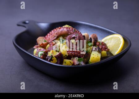 Warmer Salat mit Oktopus, Kartoffeln und Zitrone in einer schwarzen kleinen Pfanne. Nahaufnahme, schwarzer Hintergrund. Stockfoto