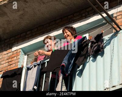 Zwei fröhliche Frauen sprechen, stehen auf dem Balkon, nachdem sie an einem sonnigen Tag nasse Kleidung zum Trocknen aufgehängt haben. Stockfoto