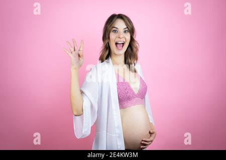 Junge schöne Brünette Frau schwanger erwarten Baby über isoliert rosa Hintergrund zeigt und zeigt mit den Fingern Nummer vier beim Lächeln CO Stockfoto