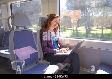 braune Haare lächelnde Frau gekleidet in lila und blau, Sitzung, die Anreise mit der Bahn, die Eingabe von Stichwort Laptop-Computer, stützte sich auf ihre Beine Stockfoto