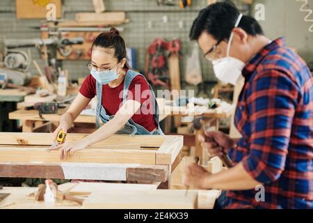 Serious junge Tischlerin in Brille und medizinische Maske Messung Holzplanken, die sie für die Herstellung von Möbelstück geschnitten Stockfoto