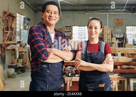 Fröhliche, reife Tischler und sein Lehrling stehen in der Werkstatt in Denim-Schürzen, klappbaren Armen und blicken in die Kamera Stockfoto