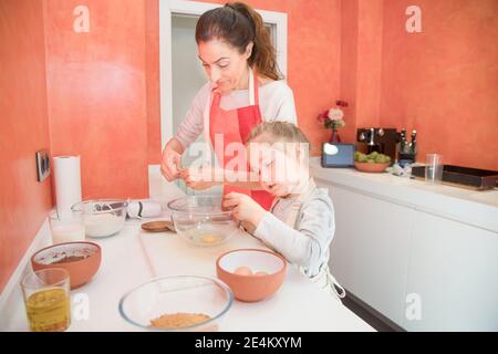 Vier Jahre altes Mädchen mit Schürze, die Eier für das Kochen von Kuchen mit der Mutter bricht, zusammen in der Teamarbeit, in der Küche zu Hause Stockfoto