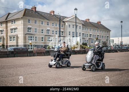 Zwei Rentner auf Mobilitäts-Scootern, die am Meer entlang fahren Hüte auf Kreuzfahrt entlang Rennen llandudno coole alte Menschen Leute nach Hause zu entkommen Stockfoto