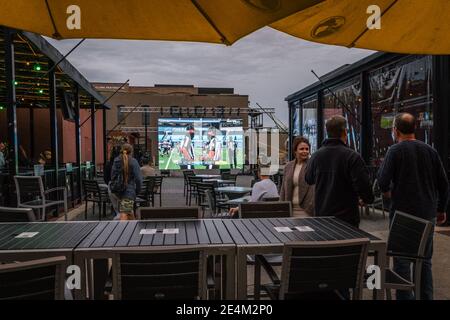 Bar mit Sitzgelegenheiten im Freien während der Covid-19 Pandemie. Gönner sind sozial distanziert & einige werden beim Betrachten eines Fußballspiels maskiert. Stockfoto