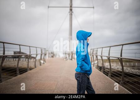 Junge in blauem Mantel mit Kapuze auf Brücke suchen Weg von der Kamera Seite Regenmantel Metall Geländer Boot Segel Hintergrund Hafen Ozean Hände in Taschen kalt Stockfoto