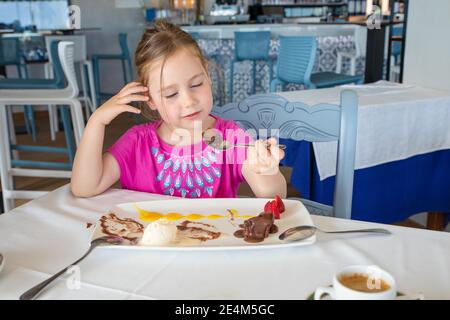 Süße vier Jahre alte blonde Mädchen essen mit Löffel süße Dessert: Schwarze Schokolade, Vanille-Eis, Erdbeeren und Mangosauce, sitzen im Restaurant Stockfoto