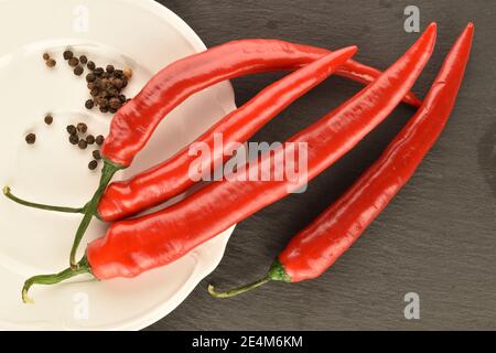 Mehrere Schoten von reifen leuchtend roten frischen Paprika auf einer weißen Keramikplatte auf einem Hintergrund einer Servierplatte aus Schiefer. Stockfoto