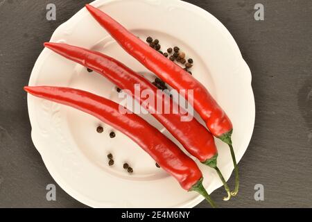 Mehrere Schoten von reifen leuchtend roten frischen Paprika auf einer weißen Keramikplatte auf einem Hintergrund einer Servierplatte aus Schiefer. Stockfoto