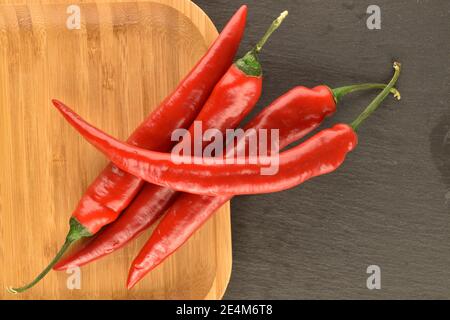 Mehrere Schoten von reifen leuchtend roten frischen Paprika auf einem Bambusblech, auf dem Hintergrund einer Servierplatte aus Schiefer. Stockfoto