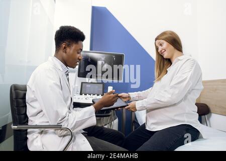 Arzt und Patient im Ultraschallraum. Afrikanischer männlicher Arzt im Gespräch mit seiner jungen schwangeren Frau Patient nach Ultraschall-Verfahren, erzählt über die Stockfoto