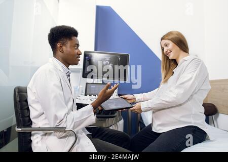 Arzt und Patient im Ultraschallraum. Afrikanischer männlicher Arzt im Gespräch mit seiner jungen schwangeren Frau Patient nach Ultraschall-Verfahren, erzählt über die Stockfoto