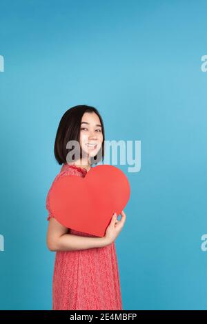 Fröhliche, fröhliche junge asiatische Frau in rotem Kleid mit großen roten Papier Herz wie Fan isoliert auf blauem Hintergrund Stockfoto