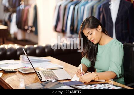 Schöne junge indische Mode Atelierbesitzer sitzen am Schreibtisch mit Laptop geöffnet und im Planer schreiben Stockfoto