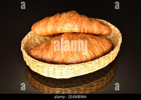 Auf einem Strohteller liegen zwei ganze duftende, frische, knackige Croissants auf schwarzem Hintergrund. Stockfoto