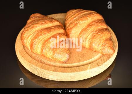 Zwei ganze duftende süße frische knusprige Croissants auf schwarzem Hintergrund liegen auf einem Holztablett Stockfoto