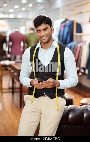 Portrait von lächelnd gut aussehend junge indische Schneider mit Anwendung auf Smartphone im Atelier Stockfoto