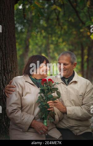 Ein Paar Rentner sitzt in einem Herbstpark auf einer Bank, ein Mann gibt einer Frau Blumen. Hochwertige Fotos Stockfoto