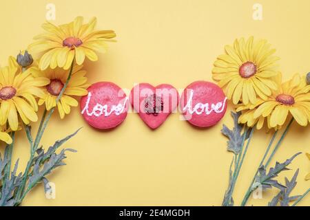 Drei rote Macarons mit Liebe in Glasur und einer geschrieben Herz geformt auf gelbem Hintergrund mit frühlingsgelben Gänseblümchen Blumen mit Kopierer Platz und Raum für Stockfoto