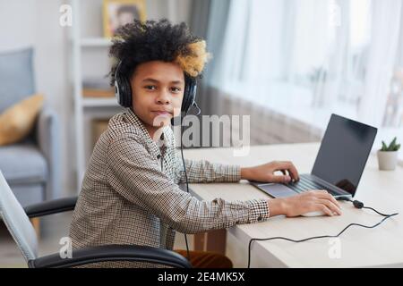 Porträt eines afroamerikanischen Jungen im Teenageralter, der Headset trägt und Kamera ansieht, während er zu Hause einen Laptop benutzt, ein Konzept für junge Gamer oder Blogger, Kopierraum Stockfoto