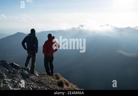 Zwei Männer stehen mit Trekkingstöcken am Klippenrand und schauen auf Sonnenuntergangsstrahlen über den Wolken. Erfolgreiches Summit-Konzept-Image. Stockfoto