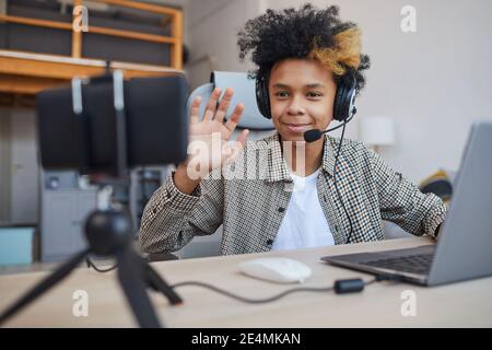 Porträt eines afroamerikanischen Jungen im Teenageralter, der Headset trägt und bei der Kamera winkt, während er zu Hause Videospiele streamt, junger Gamer oder Blogger-Konzept, Cop Stockfoto