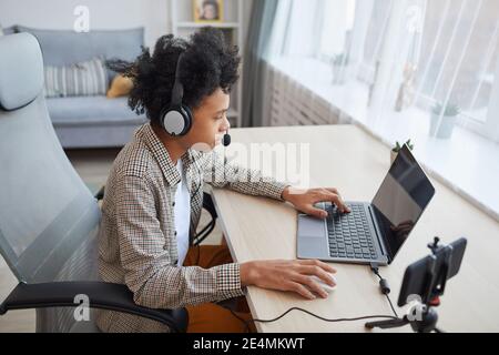 Hochwinkelporträt eines afroamerikanischen Jungen im Teenageralter, der ein Headset mit einem Laptop trägt, während er Videospiele zu Hause streamt, ein Konzept für junge Gamer oder Blogger, Stockfoto