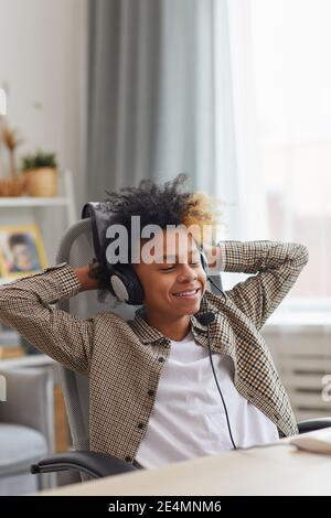 Vertikales Porträt eines lächelnden afroamerikanischen Jungen, der Headset trägt und sich im Stuhl entspannt, während er zu Hause einen Laptop, ein Gamer- oder Blogger-Konzept verwendet Stockfoto