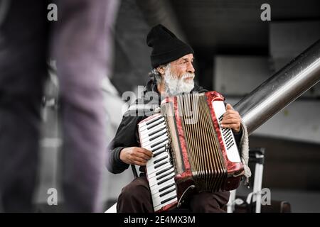 Alter Mann mit Bart spielt Straßenmusik Akkordeon trägt ein hut mit Passanten für Geld in der Stadt auf Kälte Tag unterhaltsam Busker verwittert leben Stockfoto