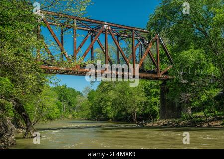 Ein sich schnell bewegender Fluss, der unter dem alten rostigen Eisen fließt Zugtreste mit Wäldern umgeben auf einem lebendigen blauen Himmel Tag im Frühling Stockfoto